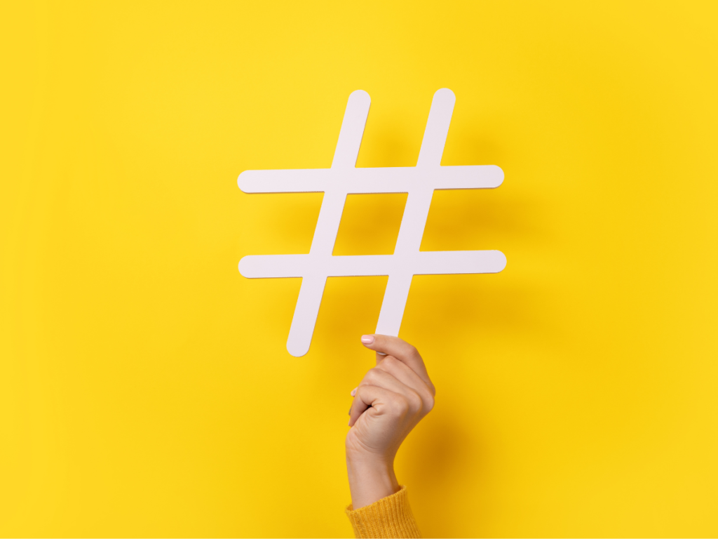 A white hashtag being held by a hand in front of a yellow backdrop.